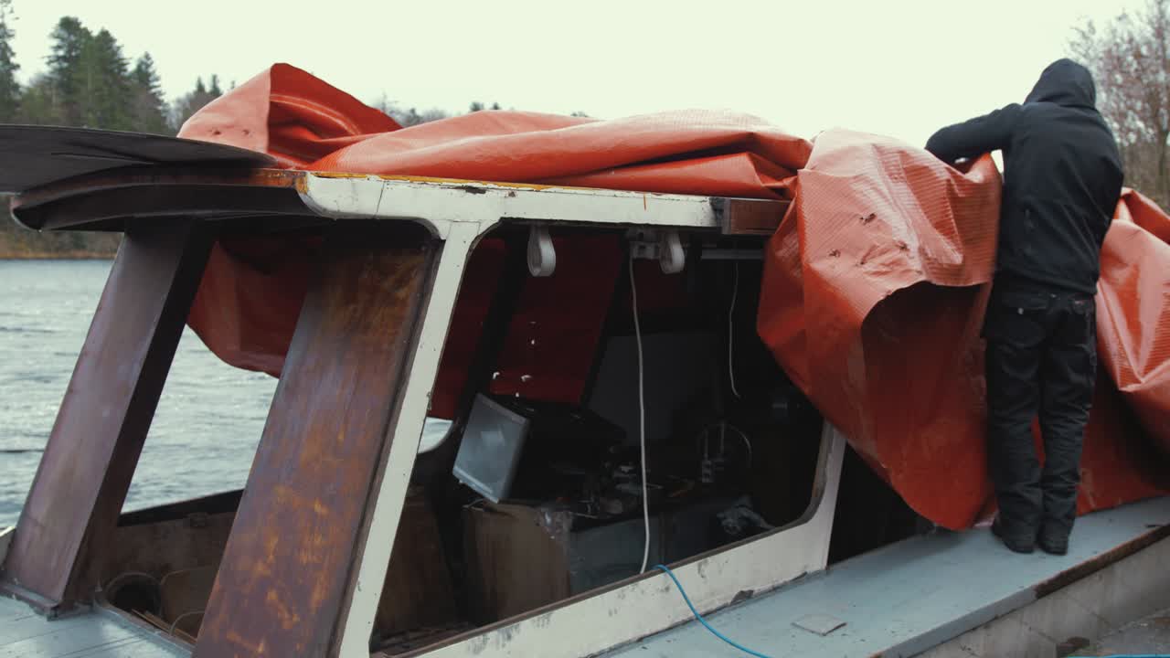 Young man untying waterproof cover of wooden boat before starting restoration maintenance