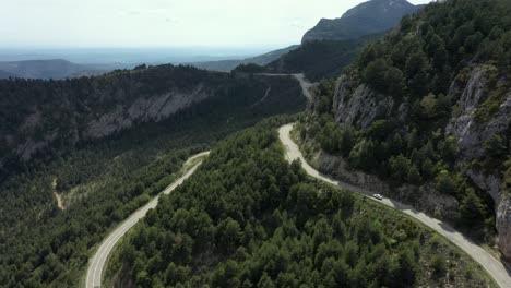 Aerial-view-small-white-car-along-curving-scenic-mountain-woodland-empty-road-into-distance