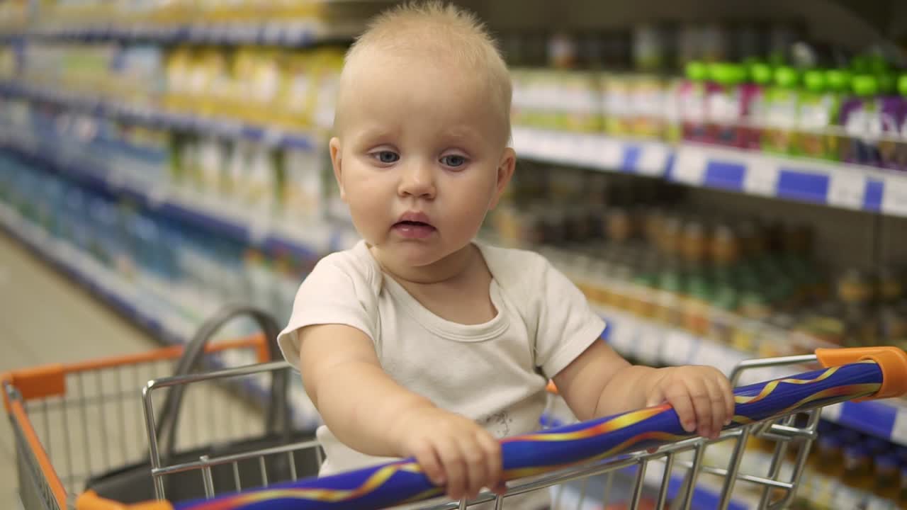 pequeño bebé sentado en un carrito de comestibles en un supermercado, mientras que su madre está eligiendo productos. compras familiares con un poco