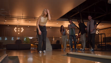 a woman in a bowling alley throws a ball on the track and knocks out a shot in slow motion and jumps and dances joy. friends support and clap with a smile. multi-ethnic friends play bowling together.