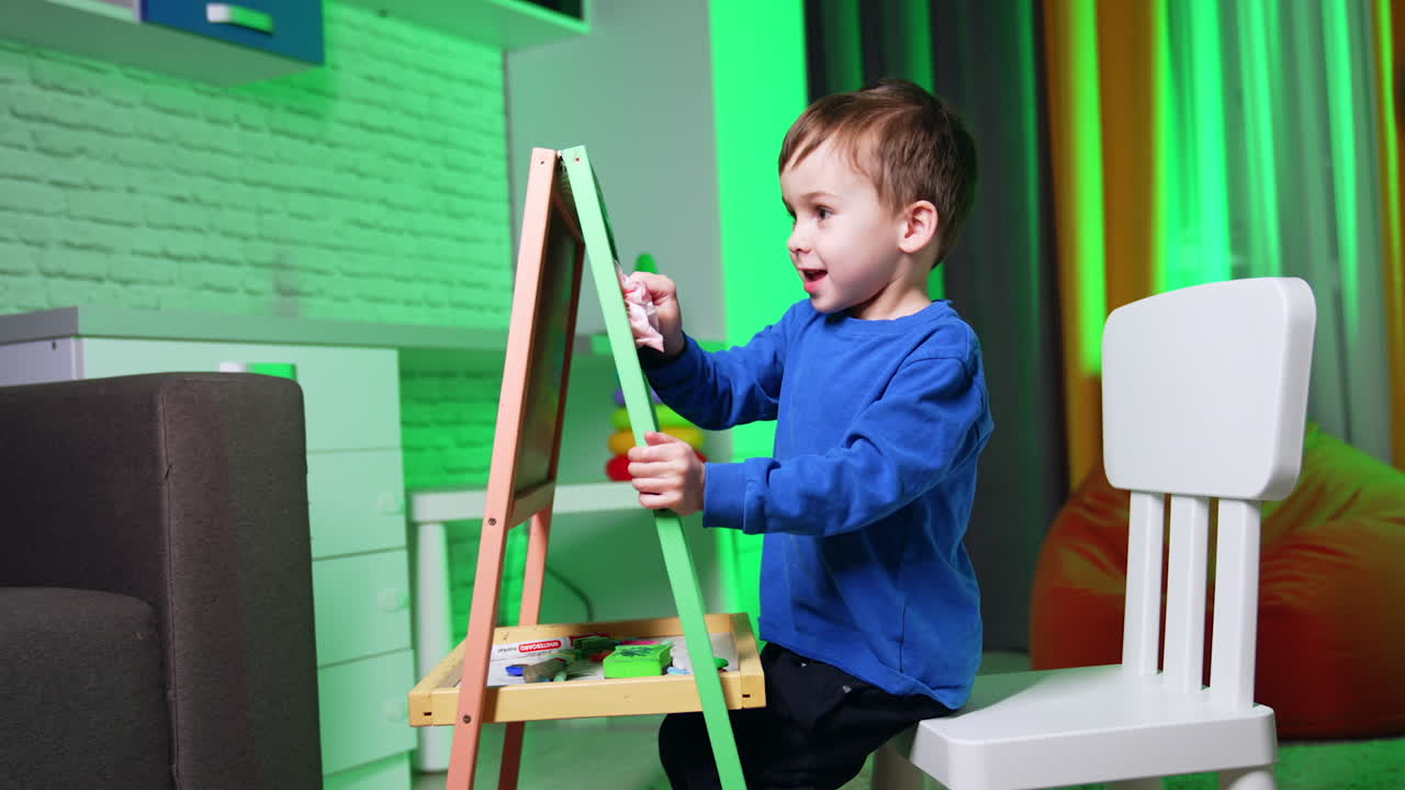 Small toddler kid sitting at the blackboard cleans it with a napkin. Baby boy takes a chalk and draws on the board.