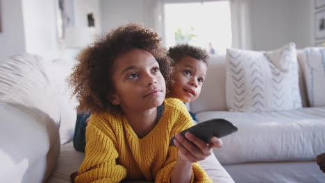 Close-up-of-pre-teen-African-American-girl-lying-on-sofa-watching-TV,-her-younger-brother-lying-behind-her