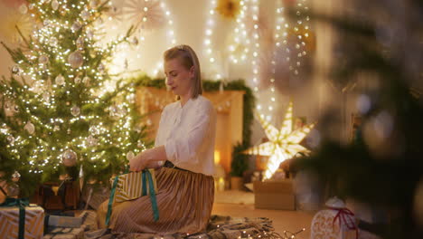 woman sits by the christmas tree and wraps christmas presents in a cozy apartment