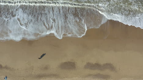 Toma-Aérea-De-Una-Pareja-Con-Sombras-Alargadas-De-Pie-En-Una-Playa-De-Arena-Dorada-Y-Admirando-El-Mar