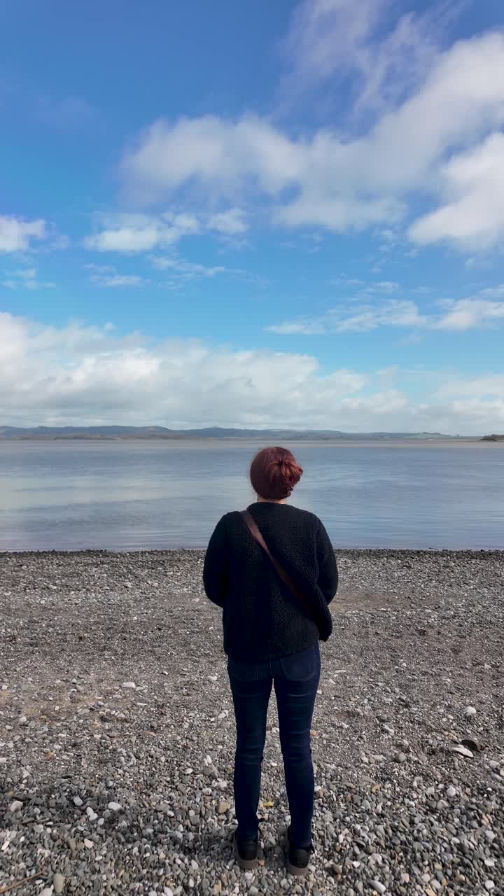 Woman standing on pebble beach looking at the calm water and horizon, finding solitude. vertical video