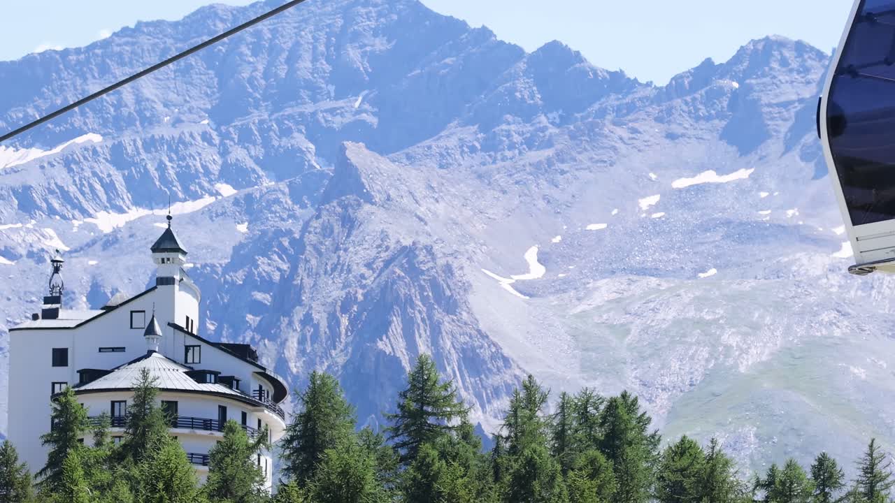 A gondola glides past a picturesque alpine building with stunning mountain peaks in the background.