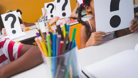 diverse class of elementary schoolchildren at their desks holding question mark signs