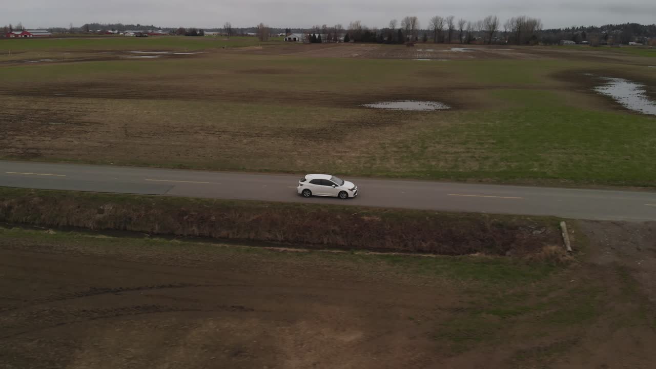 carro branco 2019 toyota corolla hatchback dirigindo ao longo da estrada rural cercada por grama campos marrons montanhas em terras agrícolas abbotsford bc carro de rastreamento líder aéreo acelera perfil lateral