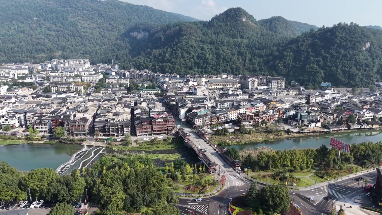 Lenta orbita aerea su una cittadina fluviale con un ponte coperto che attraversa il fiume sottostante, edifici bassi e densi che si estendono verso montagne carsiche boscose, Zhangjiajie, Hunan, Cina