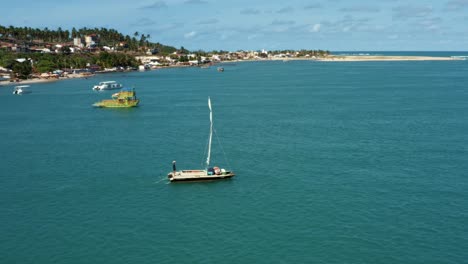 Aerial-drone-shot-following-a-small-sail-boat-boat-sailing-on-a-large-tropical-turquoise-river-to-the-Restinga-beach-from-the-Barra-do-Cunhaú-beach-in-Rio-Grande-do-Norte,-Brazil