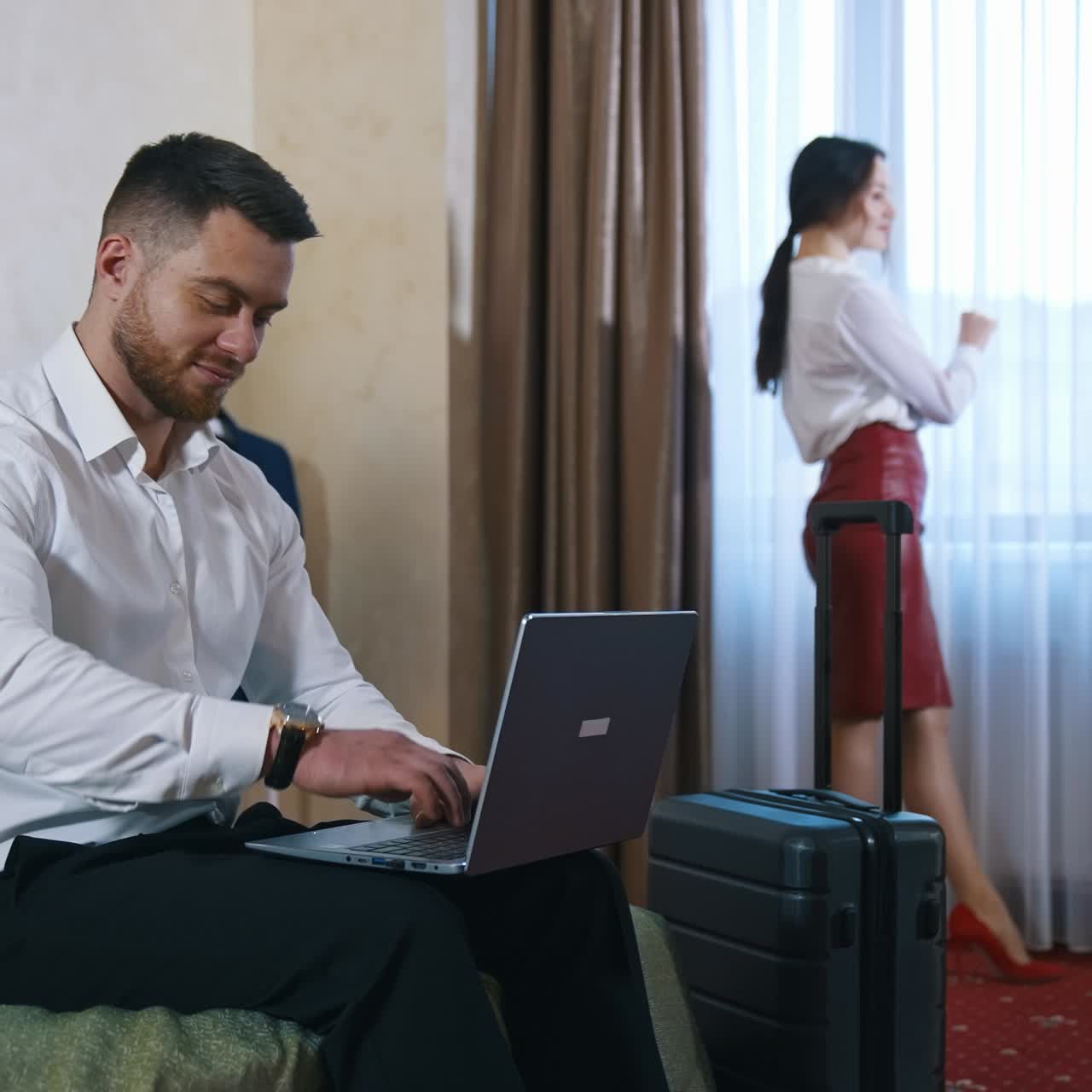 Business colleagues in a hotel room. Handsome man sitting on a bed and working on his laptop. Pretty woman in elegant clothes standing near the window in hotel