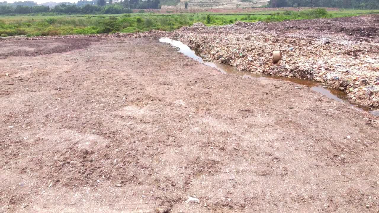Close-up shot of an enormous landfill waste pile with a channel of dark, polluted leachate liquid runoff flowing beside it, highlighting severe water contamination
