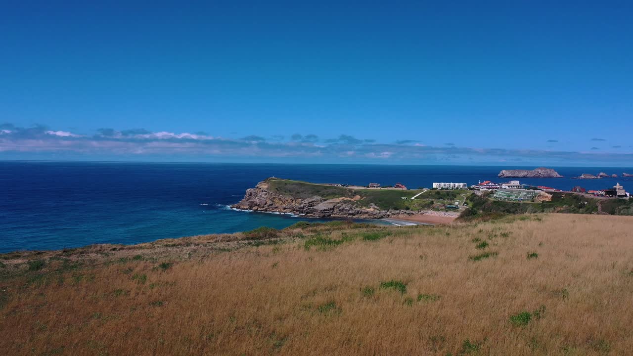 건조한 초원의 끝에서 우리는 바다로 뛰어들어 건물과 파도가 있는 해변을 발견합니다. 우리는 배경에 여름에 섬을 볼 수 있습니다. - 칸타브리아, 스페인