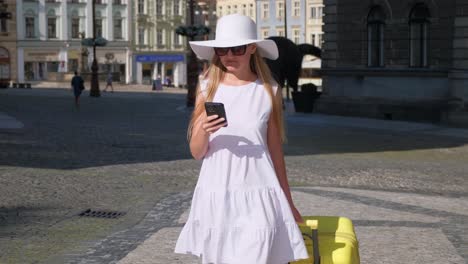 a slim beautiful woman in a white dress and hat walks with a yellow suitcase in the old city. a female tourist is heading to the hotel. summer vacation in europe