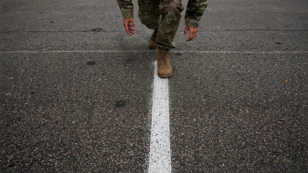Approaching person wearing camo stepping tan boots along white line on asphalt, practicing balance