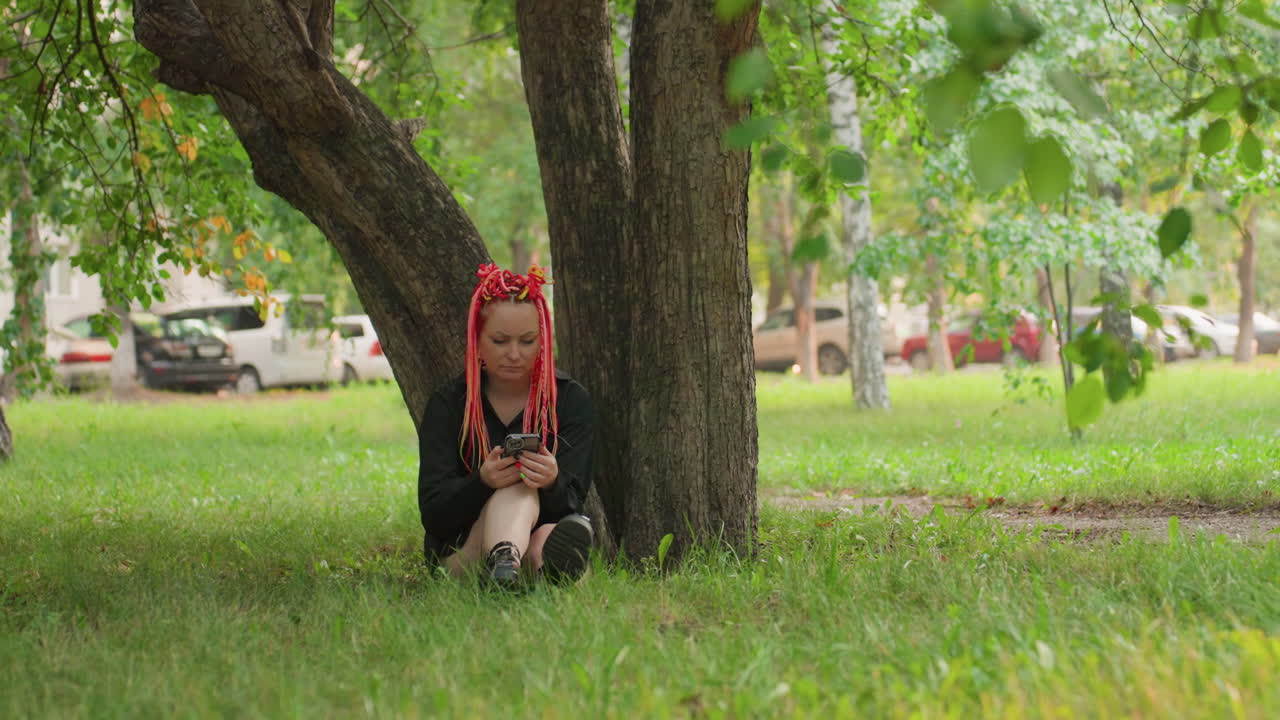 백인 십 대가 나무 아래에 앉아 휴대폰을 보고 있고, 빨간 땋은 머리, 검은색 의상, 풀밭 배경, 멀리 보이는 자동차, 조용한 오후, 엄숙한 표정, 메시지 스크롤, 사색적인 분위기, 도시 공원