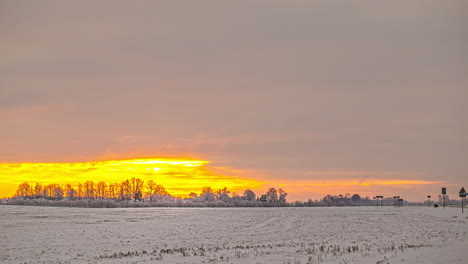 Vista-De-Los-Autos-Que-Pasan-Por-La-Carretera-Al-Lado-De-Un-Campo-Agrícola-Cubierto-De-Nieve-En-Una-Noche-Nublada-De-Invierno-En-Un-Lapso-De-Tiempo