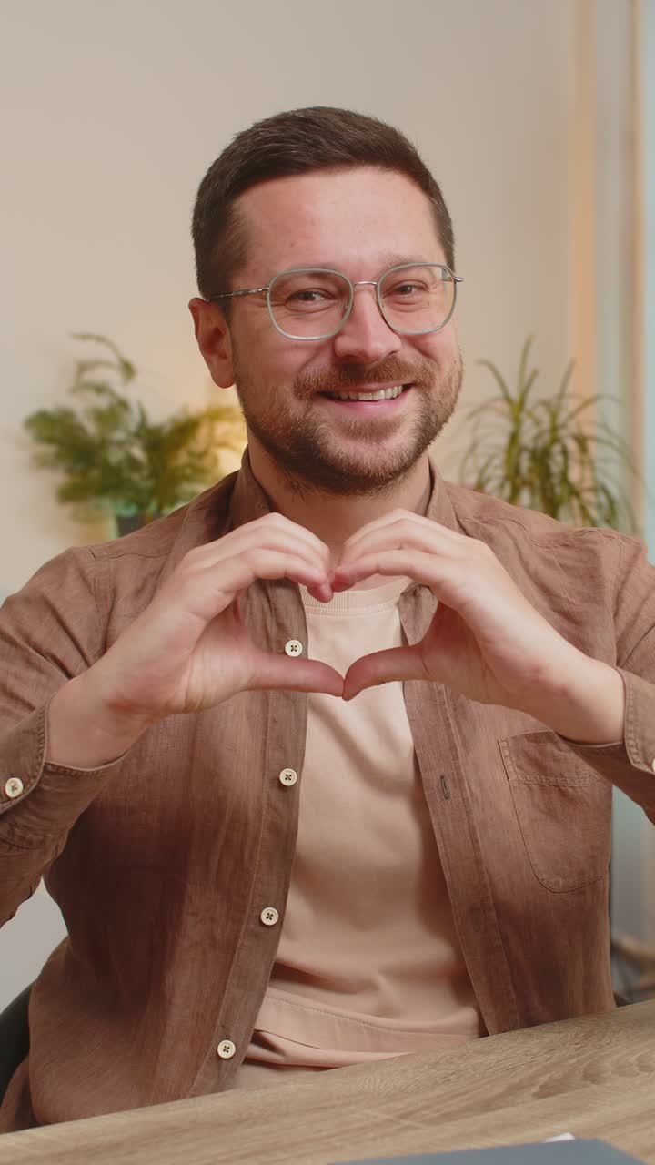 Happy caucasian young man makes symbol of love showing heart sign to camera at home office desk