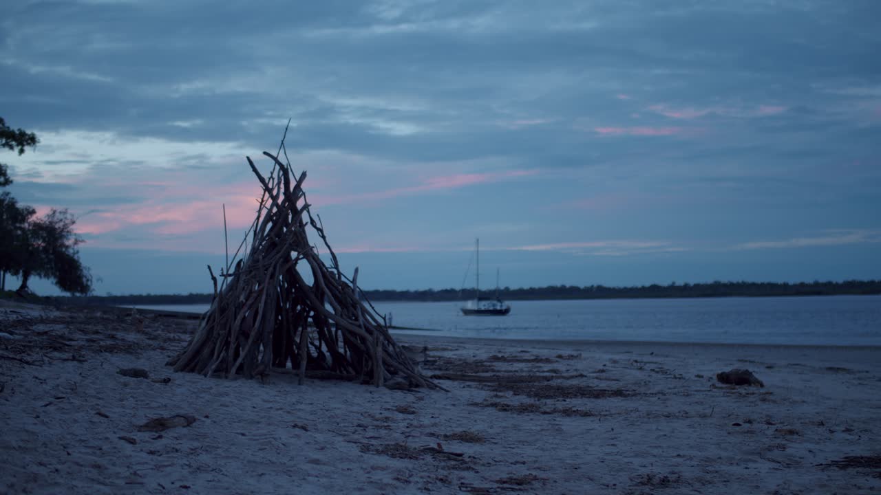 un tipi hecho de palos de varios tamaños se sienta en la tranquila playa mientras el sol en retirada derrama colores pastel de rosa y azul en las nubes, el océano y el agua mientras un bote flota a lo largo de la costa