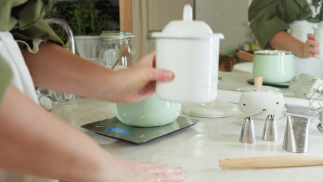 chef in apron stands near countertop after bringing down white pot, opening it and dropping lid beside as she prepares to lift pot, with kitchen utensils