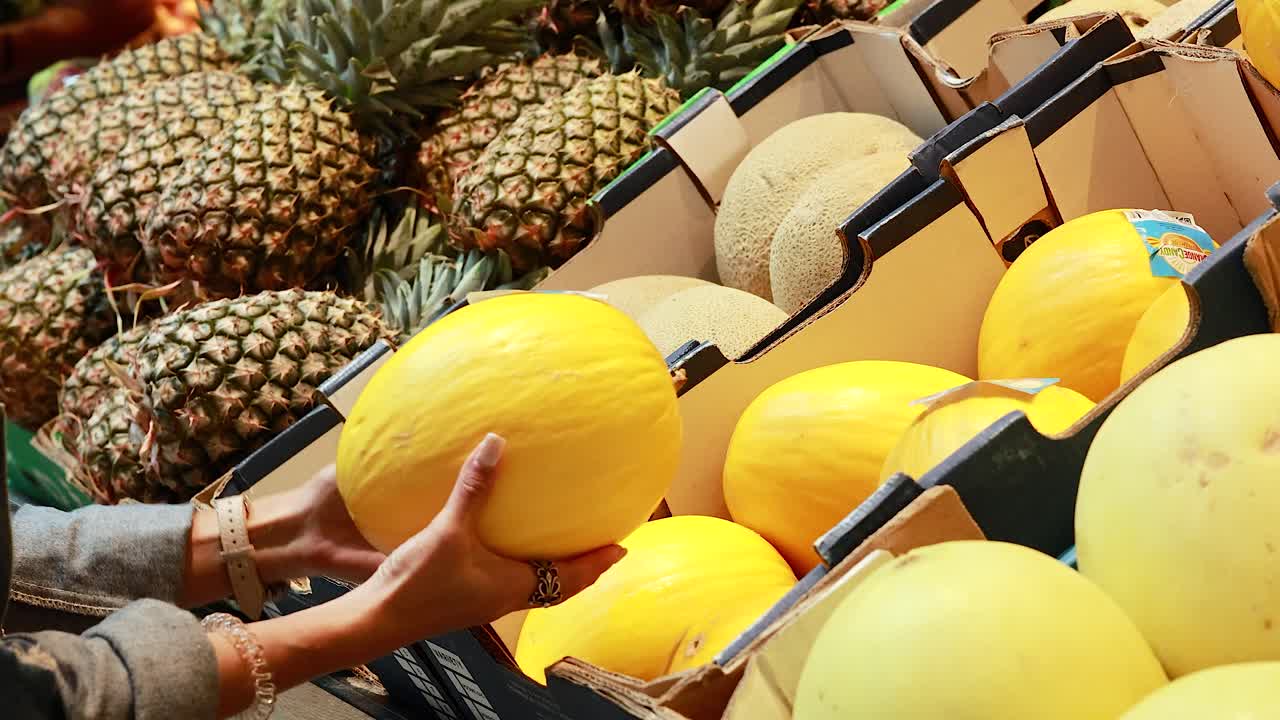 A person examines canary melons in a well-lit supermarket, surrounded by pineapples and cantaloupes, highlighting fresh produce selection