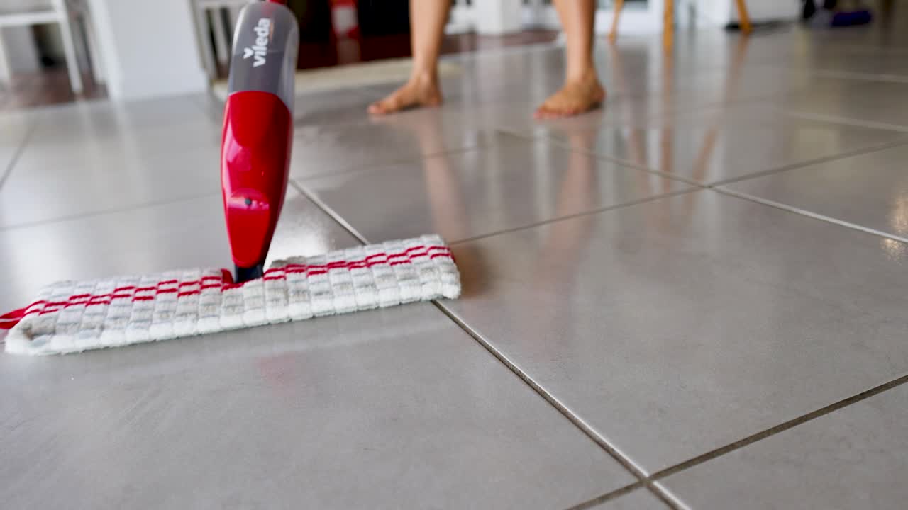 A red spray mop glides over a tiled floor, cleaning efficiently in a bright, modern room