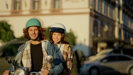 happy couple riding a motorcycle, a brunette guy with curly long hair in a denim jacket and his brunette girlfriend with long hair in a checkered shirt ride through the summer city and look at the camera