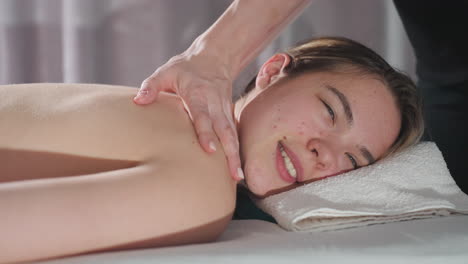 close view of massage therapist massaging client neck and shoulder while client smiles with eyes closed, light reflecting off skin creating relaxing, warm, and calming spa atmosphere