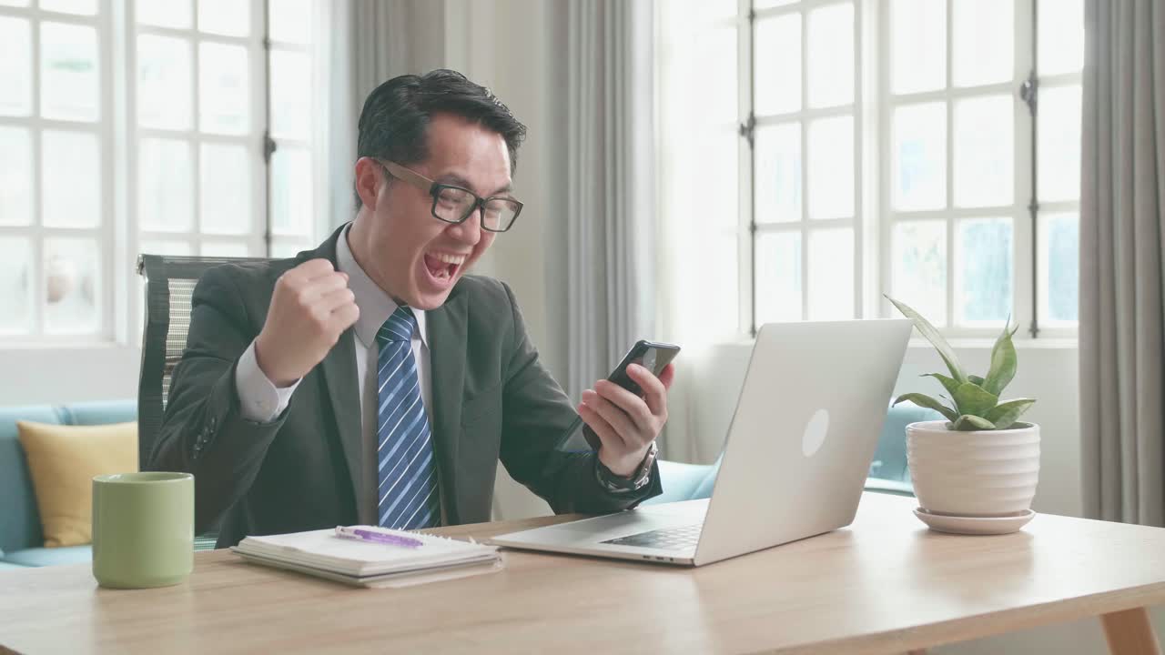 Asian Businessman With Glasses Wearing Business Suit Looking At Mobile Phone And Being Happy Winning The Project Bid While Using Computer For Working At Home.