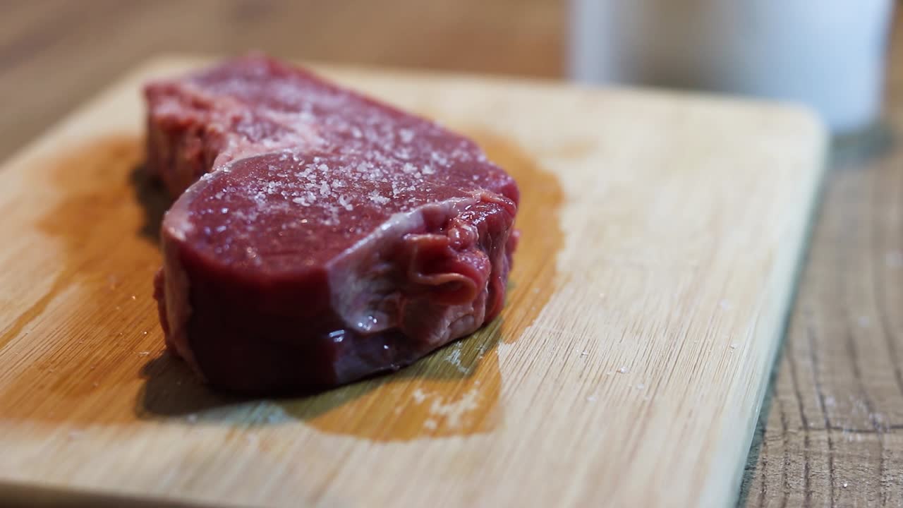 A home chef sprinkles salt over a thick beef steak resting on a wooden cutting board, preparing it for cooking on the kitchen counter.