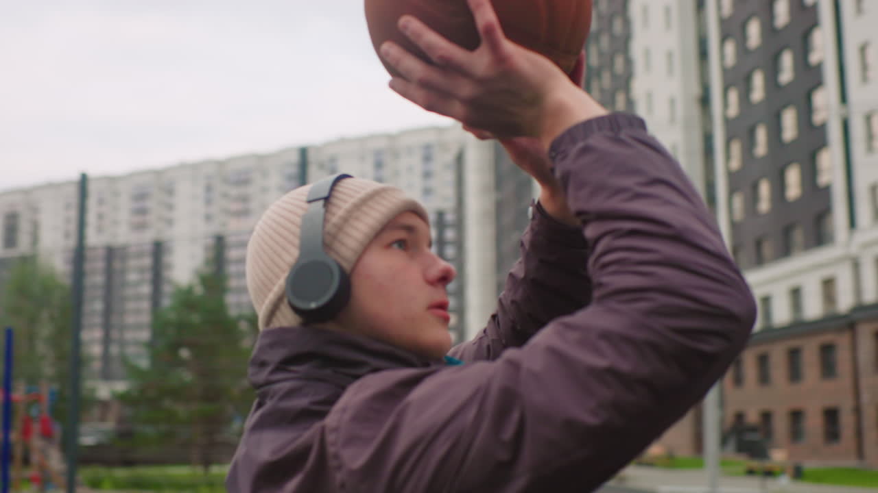 White male launching basketball at hoop beanie headphones, dynamic follow through with elevated arm and focused gaze, modern apartment skyline and park court backdrop, hopeful energy and clear arc