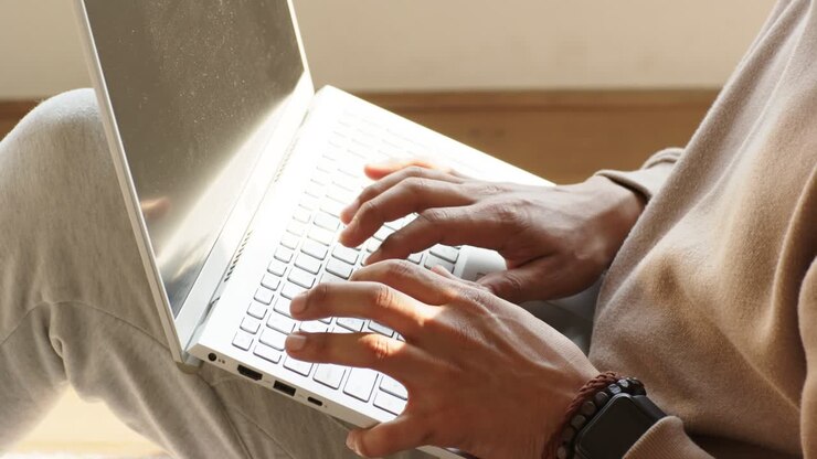 Hands sliding white laptop onto lap, typing text on keyboard above light carpet near wood wall