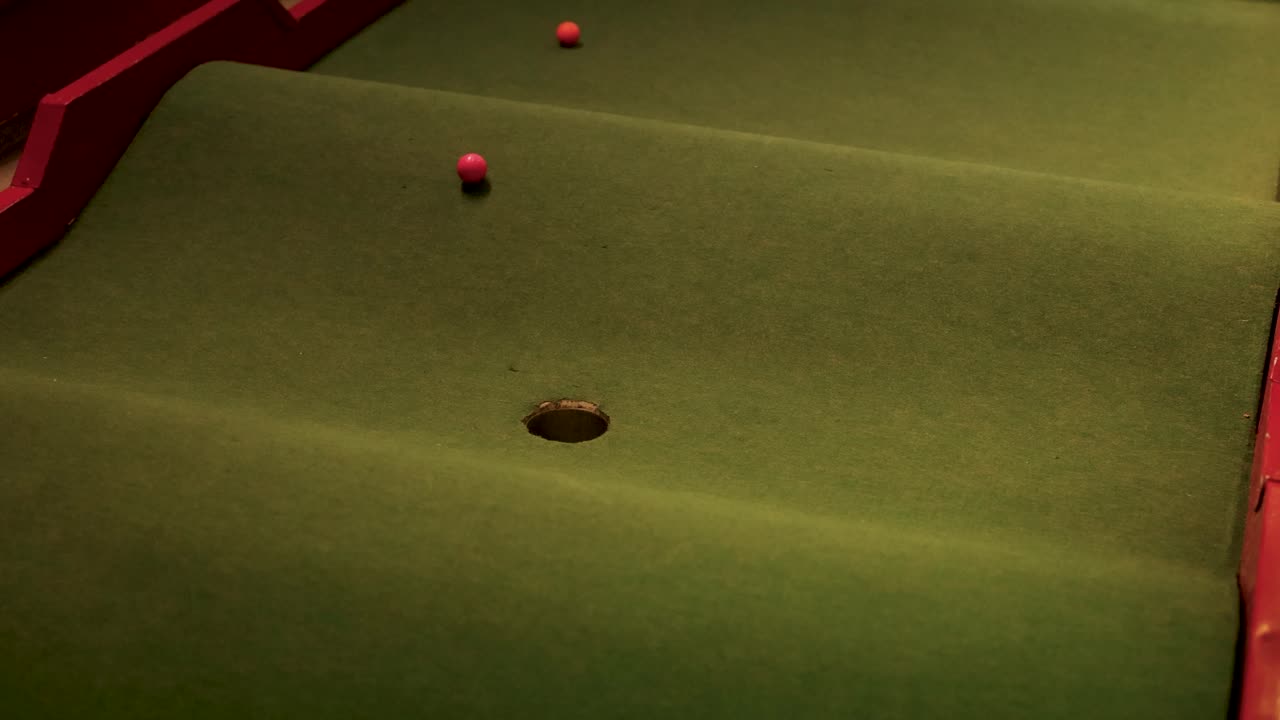 Dwie piłeczki golfowe poruszają się w przód i w tył na polu do minigolfa z dołkiem, w słabym oświetleniu.
