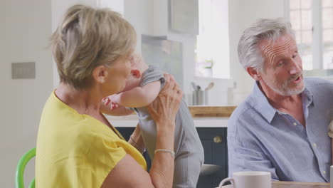 Grandparents-Looking-After-Laughing-Granddaughter-And-Baby-Grandson-Sitting-At-Kitchen-Table