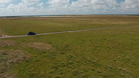 Aerial-shot-of-car-going-on-narrow-road-in-countryside.-Flat-grassland-and-forest-in-distance.-Denmark