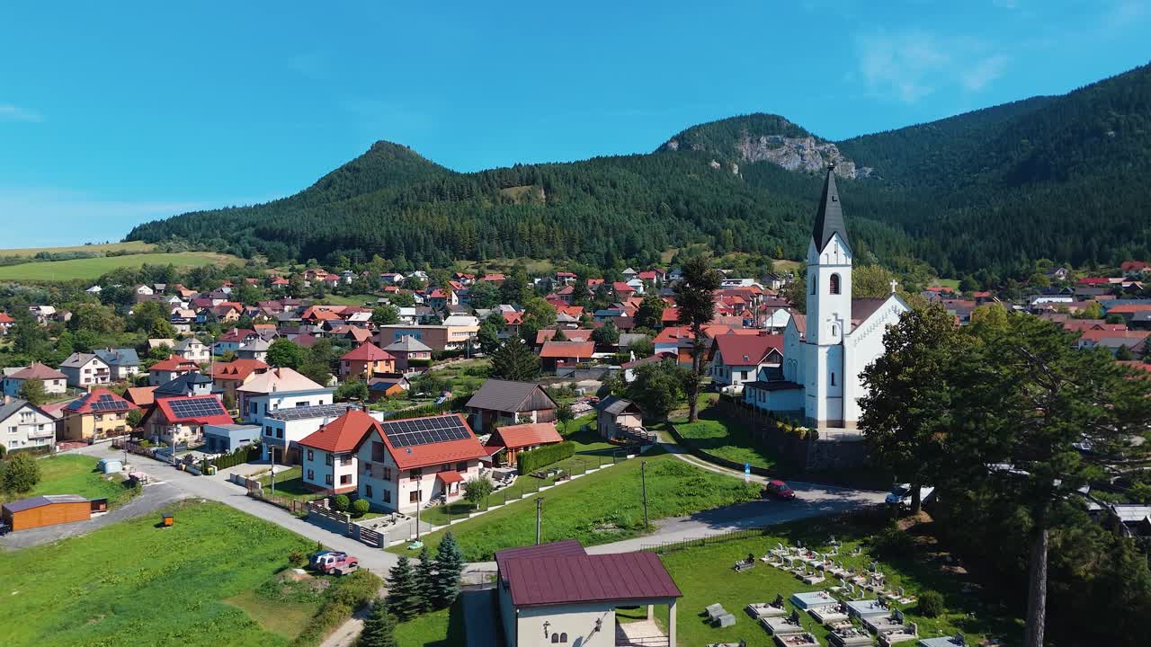 Drone etenee slovakialaisen kylän yllä, jossa on valkoinen kirkko ja punakattoisia taloja vihreiden vuorien ympäröimänä kirkkaan sinisen taivaan alla.