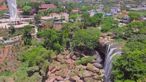 This-footage-captures-a-breathtaking-Waterfall-in-Vietnam,-cascading-down-a-rocky-cliff-with-a-beautiful-female-Buddha-visible-in-the-background