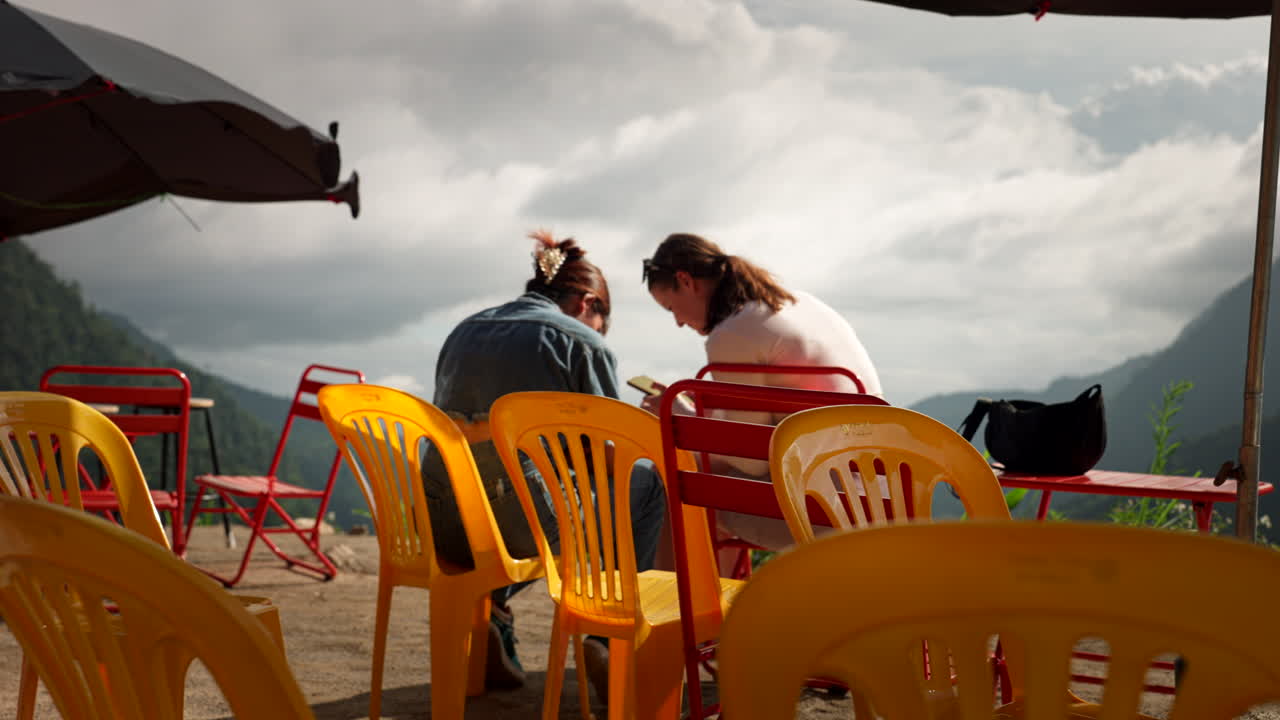 Engaging conversation between tourist and local in Ha Giang village, vibrant cultural exchange as they sit on plastic chairs, medium rearview