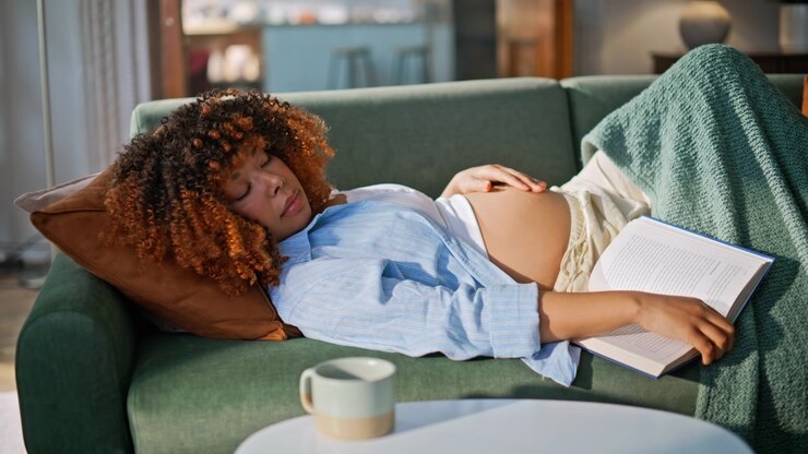 Napping pregnant woman resting sofa with book. Tired mother touching belly home.