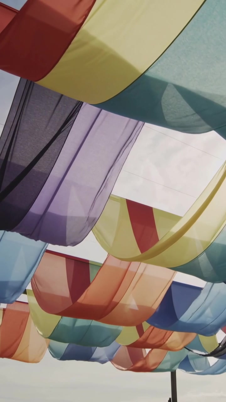 Vertical video: Breeze lifting thin panels overhead shading walkway, showing pole, blending colors