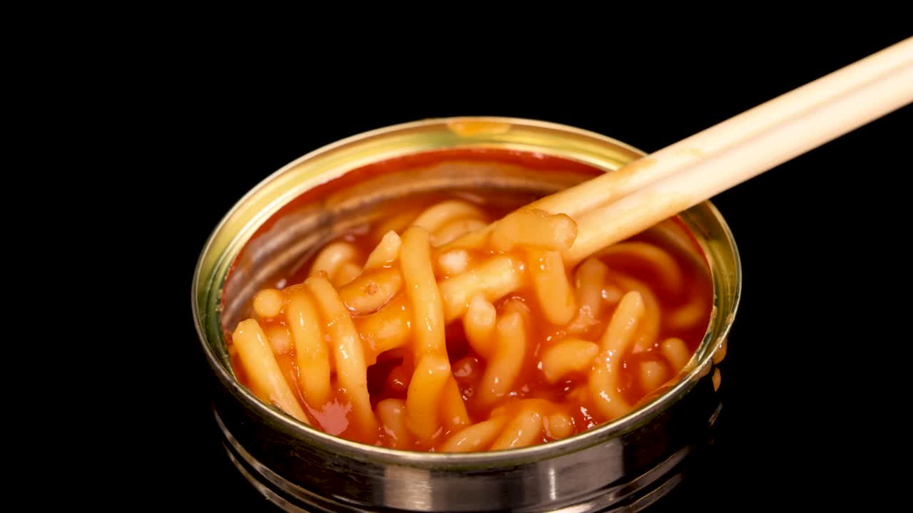 Chopsticks grasp and lift spaghetti in tomato sauce from an open metal can, isolated on a black background with even studio lighting and close-up framing