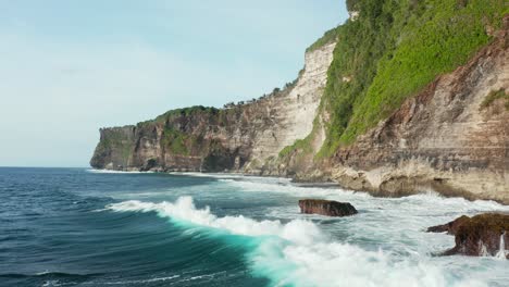 low angle drone shot flying over water next to tall seaside cliffs covered with green forest on the right side