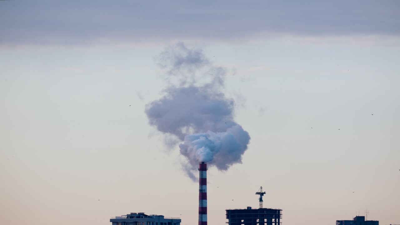 Ciminiera industriale che rilascia fumo denso nel cielo sopra un cantiere edile cittadino.