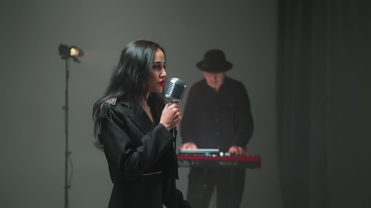 검은 옷을 입은 보컬이 빈티지 마이크에서 열정적으로 노래하고, 키보디스트는 배경에서 흐릿하게 보입니다.