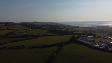 Vista-Aérea-Del-Parque-De-Caravanas-De-Gales-Al-Amanecer-Con-Vistas-A-La-Reluciente-Bahía-Costera-Y-A-Las-Onduladas-Colinas-Del-Campo,-Panorámica-A-La-Derecha