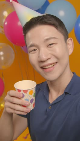 파티 모자를 입고 생일을 축하하는 남자의 수직 비디오 스튜디오 초상화는 풍선으로 종이 컵으로 카메라에 환호합니다.