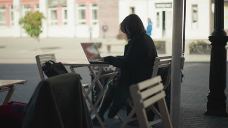 woman seated outdoors at cafe table working on laptop surrounded by wooden chairs and potted plants, with paved walkway and blurred city background