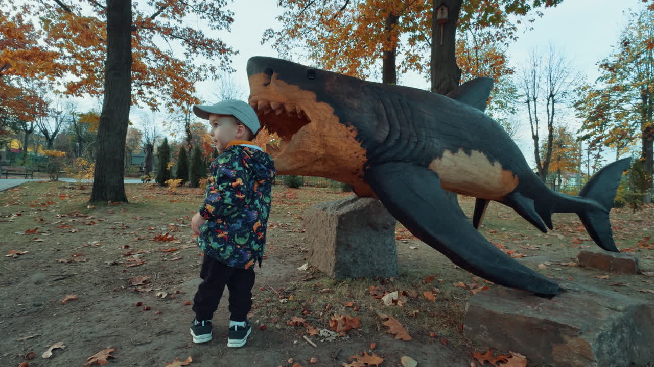 Curious toddler wearing warm clothes stands near the wooden sculpture of shark. Lovely kid in the park in autumn.