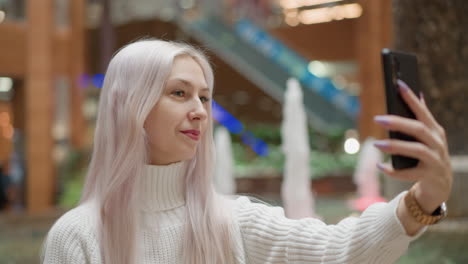 cheerful urban dweller sits on fountain bench in mall, uses phone camera for selfie series, smiling and laughing as water jets sparkle in blurred background under mall lights