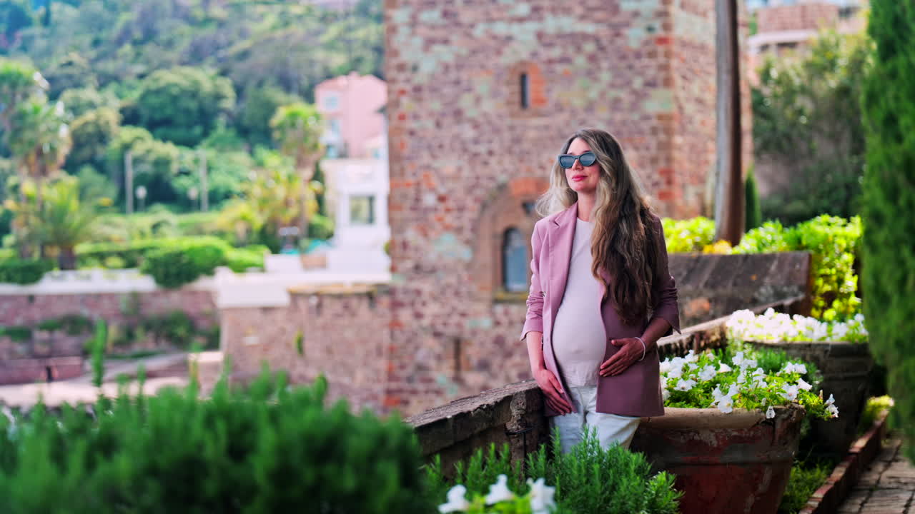 Pregnant woman with long hair, wearing sunglasses standing with the Chateau de la Napoule Castle in Mandelieu-La Napoule, France in the background